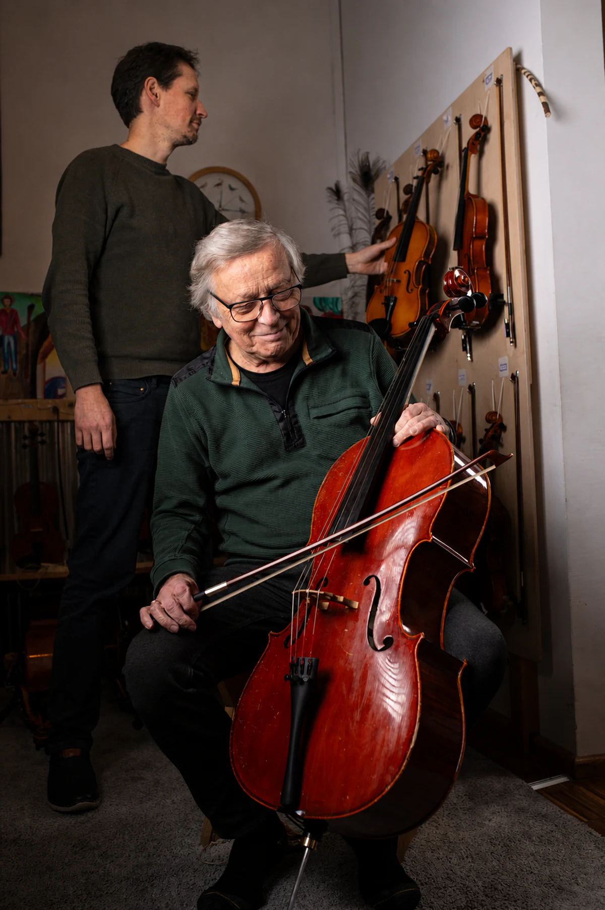 Ein Cellist sitzt mit seinem Cello vor einer Instrumentenwand, während ein weiterer Mann eine Violine betrachtet